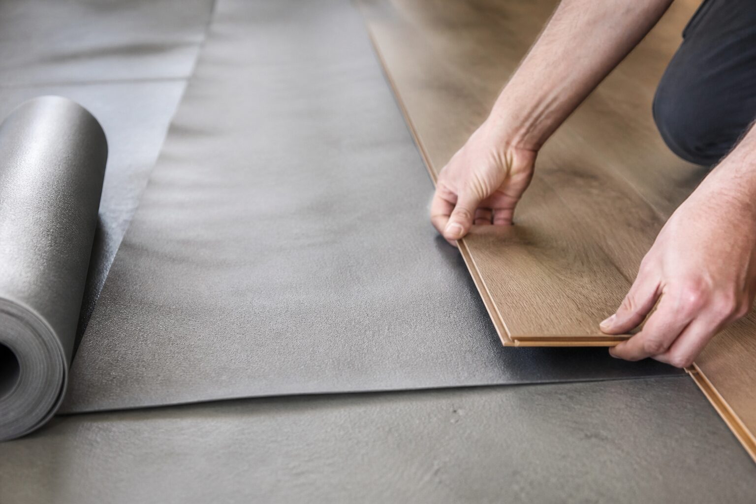 Sous-couche parquet flottant déroulée sur béton avec lame de parquet en cours d'emboîtement, mains tenant la lame, lumière naturelle